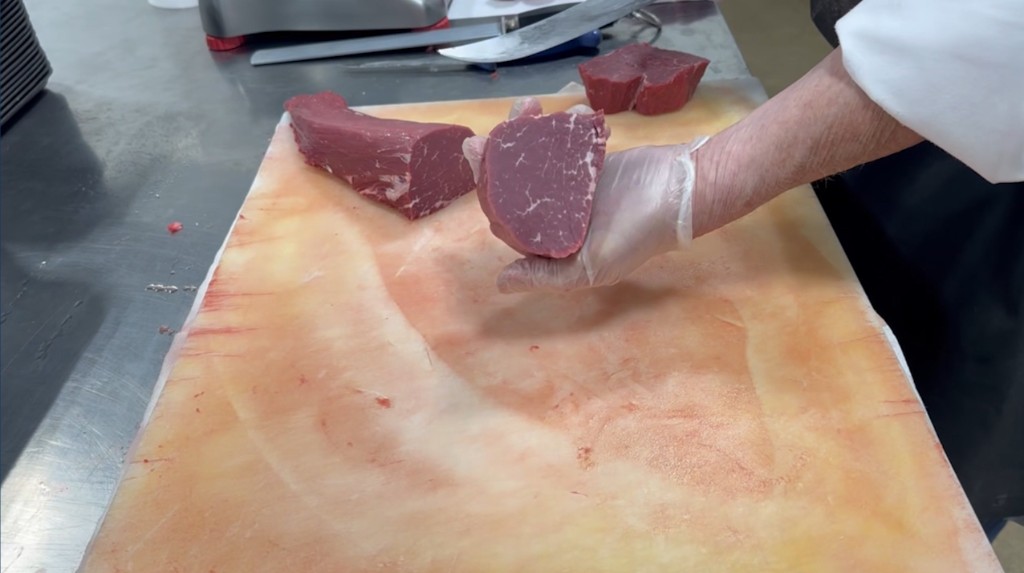 Gloved hand holding a hand-cut piece of raw meat on a cutting board